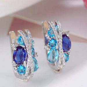 May include: A pair of silver-toned hoop earrings. Each earring features a cluster of blue gemstones in various shades, including oval and round cuts. The gemstones are accented with small, clear crystals.