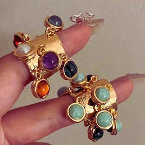 May include: Gold-toned statement rings with colorful gemstones. The rings feature a textured design and are adorned with various round and oval stones in shades of purple, green, orange, and blue. A smaller silver ring is also visible.