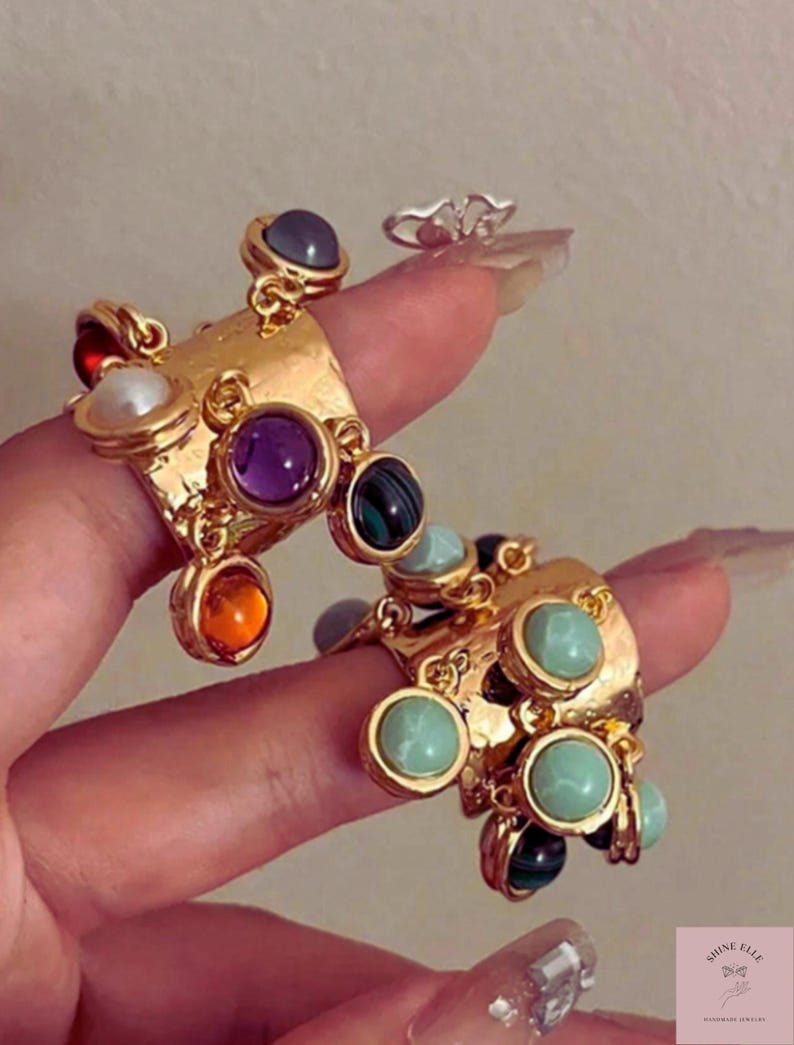 May include: Gold statement rings with colourful gemstones. The rings have a textured design and are adorned with various round and oval stones in shades of purple, green, orange, and blue. A smaller silver ring is also visible.