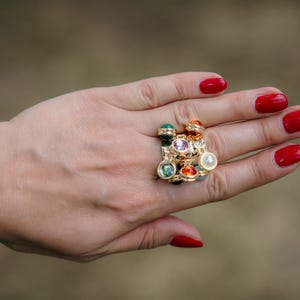 May include: A gold ring featuring an array of colourful gemstones, including green, purple, orange, and white, is shown on a hand with red nail polish. The ring has a cluster design with various shapes and sizes of stones.