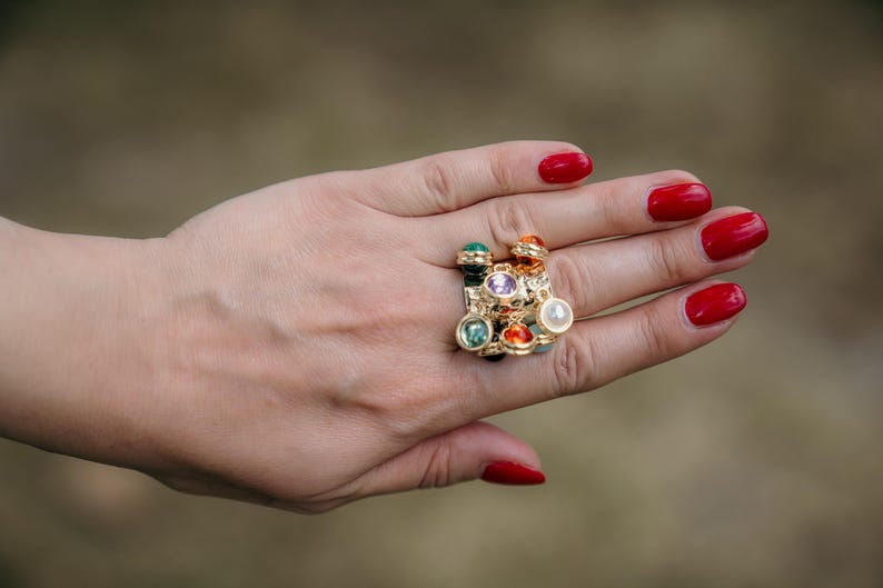 May include: A gold ring featuring an array of colourful gemstones, including green, purple, orange, and white, is shown on a hand with red nail polish. The ring has a cluster design with various shapes and sizes of stones.