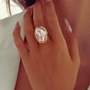 May include: A gold ring with a large, irregular, pale pink pearl. The ring's band has a textured, organic design. The jewellery is displayed on a hand, showcasing the ring's size and style. The ring is a statement piece, suitable for special occasions.