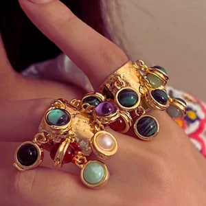 May include: Gold rings with dangling charms, each holding different gemstones. The rings have a textured design and are adorned with black, green, purple, red, and pearl-coloured stones. The image also shows a silver flower ring and the text "SHINE ELLE".