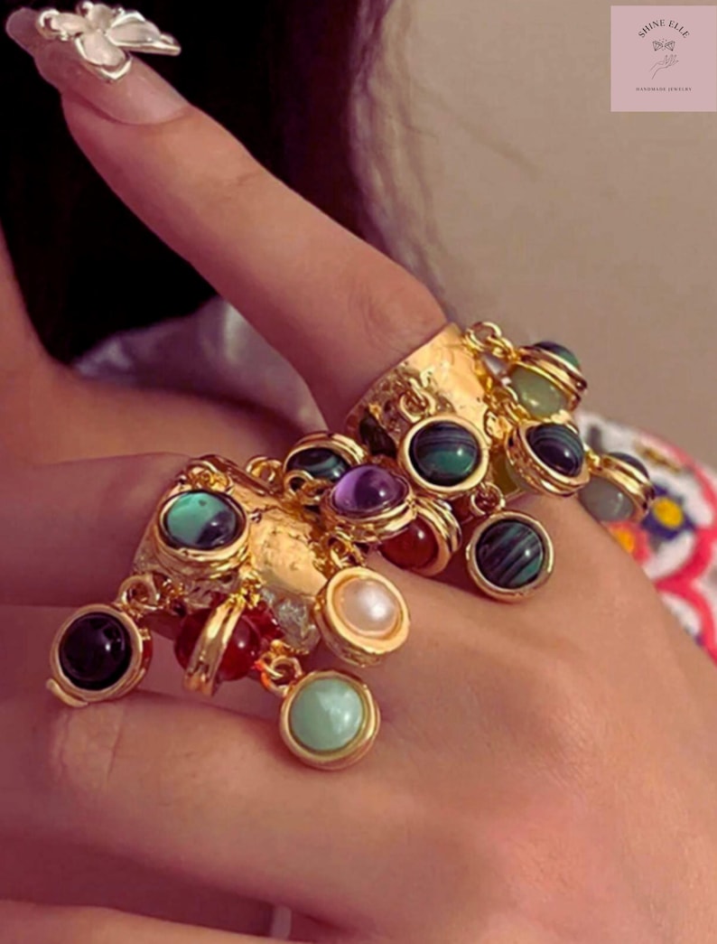 May include: Gold rings with dangling charms, each holding different gemstones. The rings have a textured design and are adorned with black, green, purple, red, and pearl-coloured stones. The image also shows a silver flower ring and the text "SHINE ELLE".