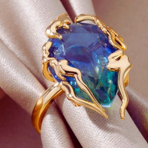 May include: A gold ring with a large, blue, faceted gemstone with green undertones. The ring has an elaborate gold setting with decorative elements. The ring is presented on a soft, light-coloured fabric. The brand name "SHINE ELLE" is visible.