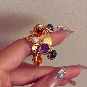 May include: A gold ring adorned with various coloured gemstones, such as red, purple, green, and orange, plus a white pearl. The ring has an elaborate design and is shown on a finger.