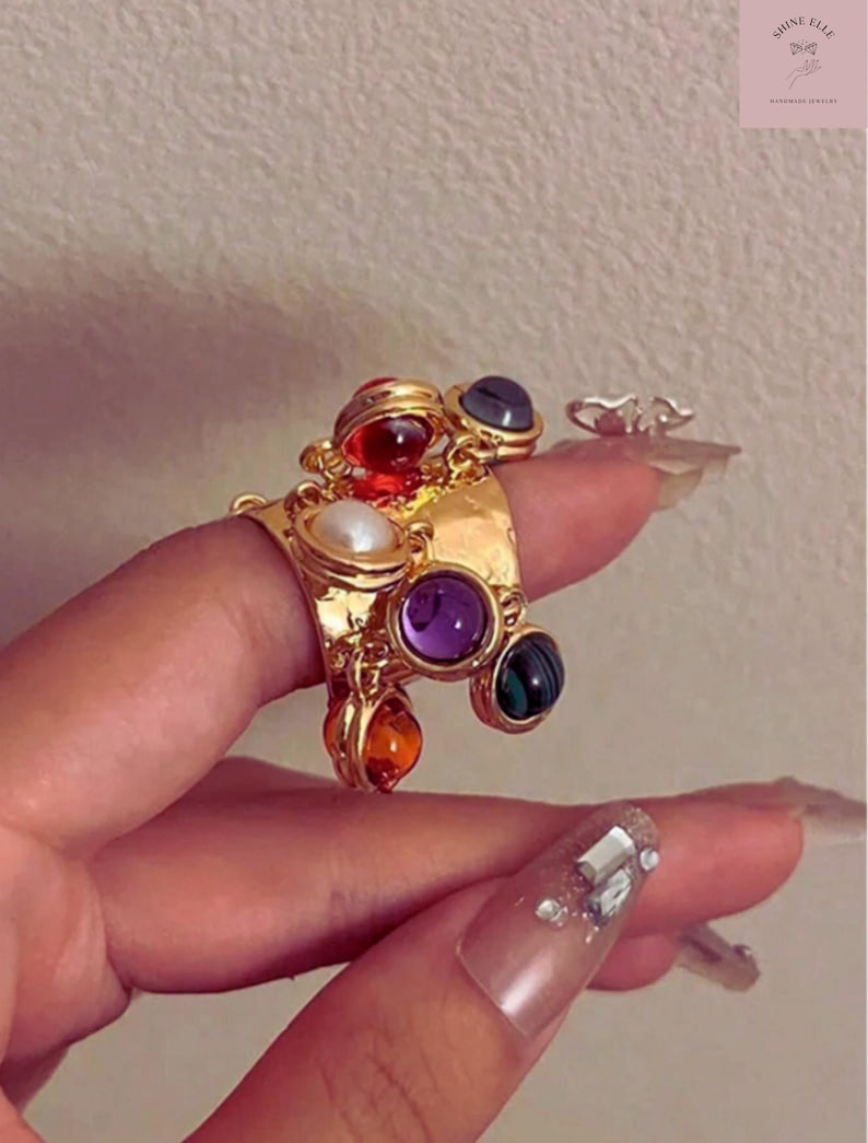 May include: A gold ring adorned with various coloured gemstones, such as red, purple, green, and orange, plus a white pearl. The ring has an elaborate design and is shown on a finger.