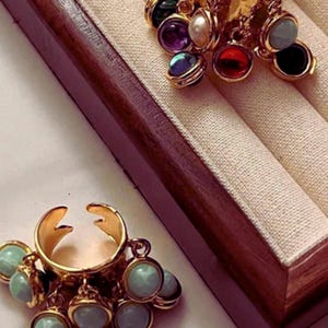 May include: Two gold rings with dangling charms. One ring has multiple colourful gemstones, the other has light blue charms. The rings are displayed on a beige fabric within a wooden jewellery box.