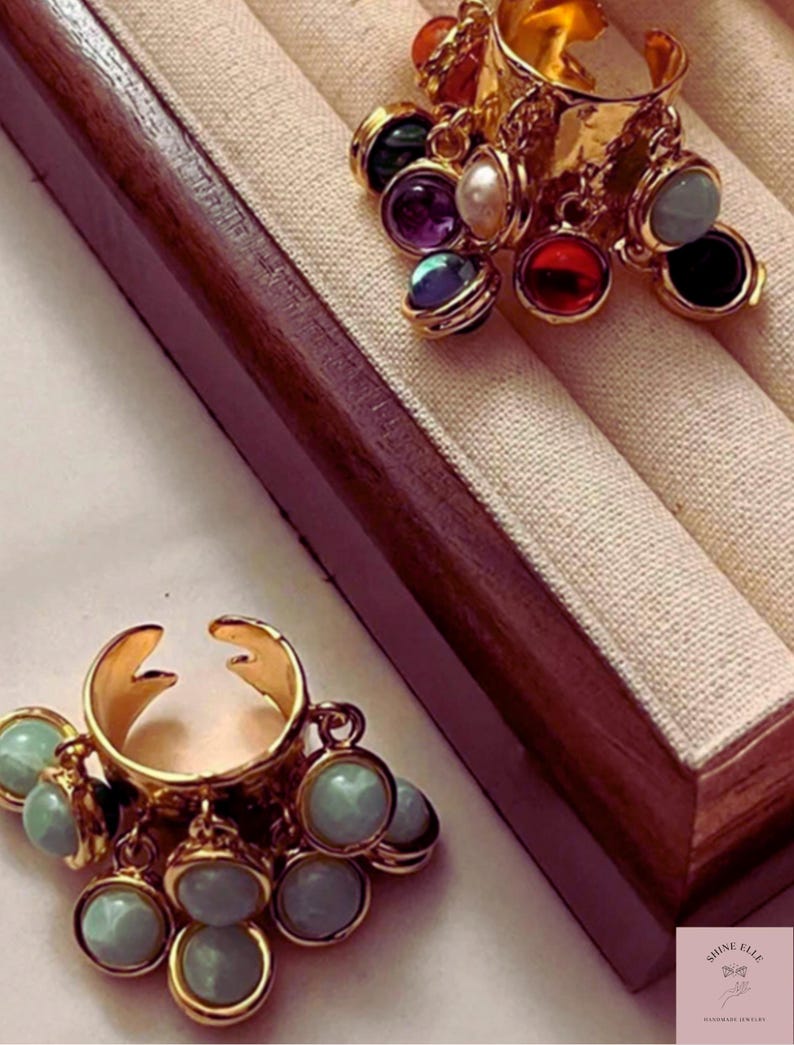 May include: Two gold rings with dangling charms. One ring has multiple colourful gemstones, the other has light blue charms. The rings are displayed on a beige fabric within a wooden jewellery box.
