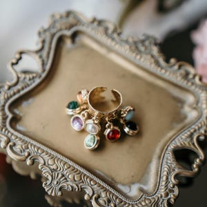 May include: A gold-coloured adjustable ring with dangling charms in various colours, including green, purple, red, and blue. The ring is displayed on a decorative gold-coloured tray with ornate edges, suggesting a vintage style.