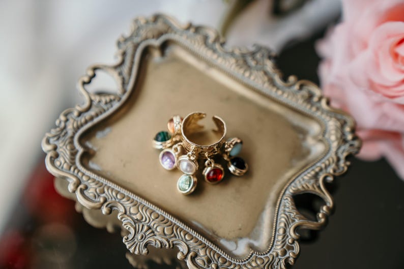 May include: A gold-coloured adjustable ring with dangling charms in various colours, including green, purple, red, and blue. The ring is displayed on a decorative gold-coloured tray with ornate edges, suggesting a vintage style.