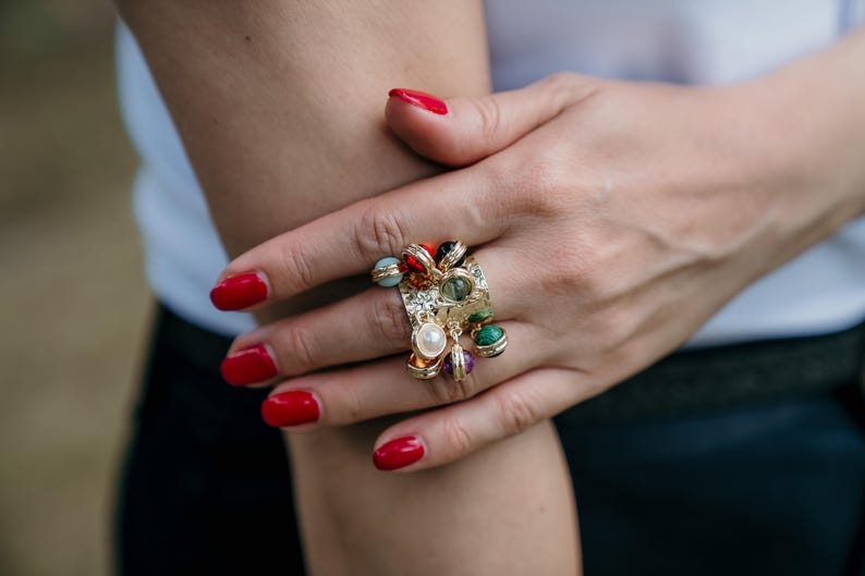 May include: A gold ring with a rectangular setting, adorned with a cluster of colourful gemstones. The ring features red, green, and pearl-like beads. The person's nails are painted red.