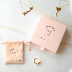 May include: A jewellery product shot featuring a gold ring, necklaces, a pink box, and a drawstring pouch. The box and pouch are branded with the text "SHINE ELLE HANDMADE JEWELRY". The ring has a floral design.