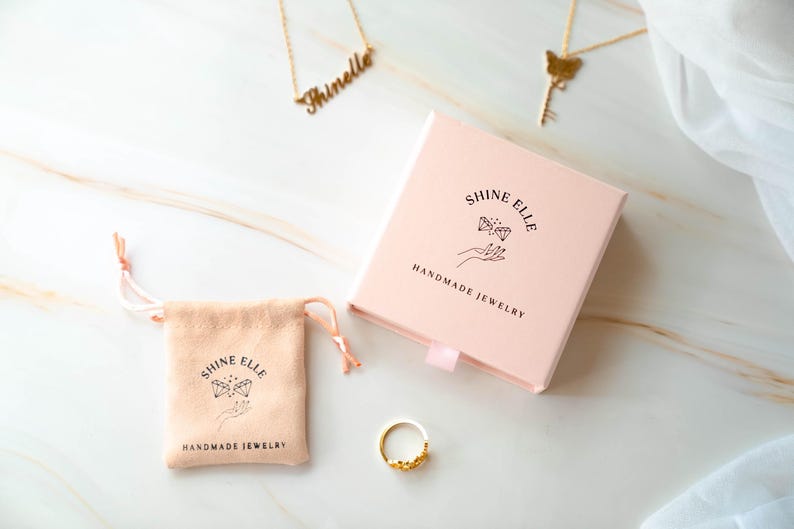 May include: A jewellery product shot featuring a gold ring, necklaces, a pink box, and a drawstring pouch. The box and pouch are branded with the text "SHINE ELLE HANDMADE JEWELRY". The ring has a floral design.