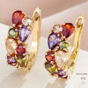 May include: A pair of gold-toned hoop earrings adorned with an array of colorful gemstones. The earrings feature a cluster of pear-shaped and round stones in shades of red, purple, green, and clear, creating a vibrant and eye-catching design.