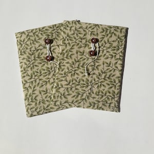 May include: Two fabric pouches with a beige background and a green leaf pattern. Each pouch has a brown button closure with white string. The pouches are folded and appear to be handmade.