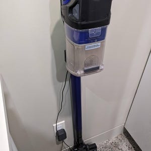May include: A blue and black Shark vacuum cleaner mounted on a wall. The vacuum has a clear dustbin filled with debris, a blue handle, and a black head. The vacuum is plugged into a wall outlet with a black charger.