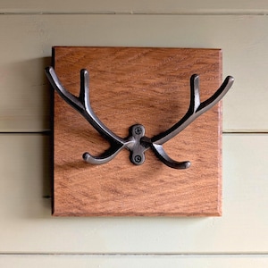 May include: A wall-mounted hook featuring a dark metallic antler design attached to a square, medium-toned wooden base. The metal fixture has two main upward-curving prongs and two smaller downward-curving prongs, secured by two visible screws.