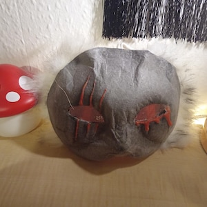 May include: A red and white mushroom-shaped lamp sits beside a grey, rock-shaped cushion with red embroidered details. The lamp has a white base. The cushion has two red embroidered shapes.