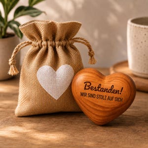Könnte beinhalten: Ein kleiner Geschenkbeutel aus Jute mit einem weißen Herzdesign neben einem herzförmigen Holzornament. Das Holzherz trägt die Inschrift "Bestanden!" und "WIR SIND STOLZ AUF DICH". Der Beutel hat einen Kordelzugverschluss.
