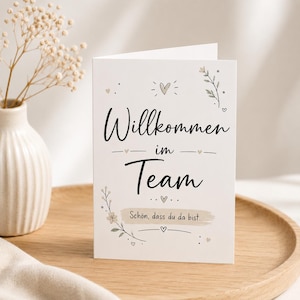 Könnte beinhalten: Eine weiße Grußkarte mit schwarzem Schreibschrift-Text, der Willkommen im Team lautet, und kleinerem Text, der Schön, dass du da bist lautet. Die Karte zeigt zarte Illustrationen von kleinen Herzen und botanischen Zweigen in gedeckten Beige- und Grautönen.