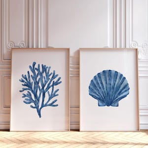Op de afbeelding: Twee ingelijste aquarelprints. Eén toont een blauw koraalontwerp, de andere een blauwe zeeschelp. Beide staan tegen een witte achtergrond met een houten frame. De prints worden op een houten vloer getoond.