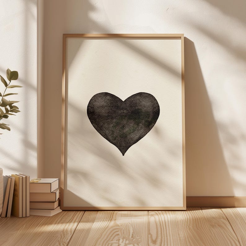 May include: A framed art print featuring a large, black watercolor heart on a cream-colored background. The artwork is displayed in a light-colored wooden frame, leaning against a wall. The image is lit by natural light.