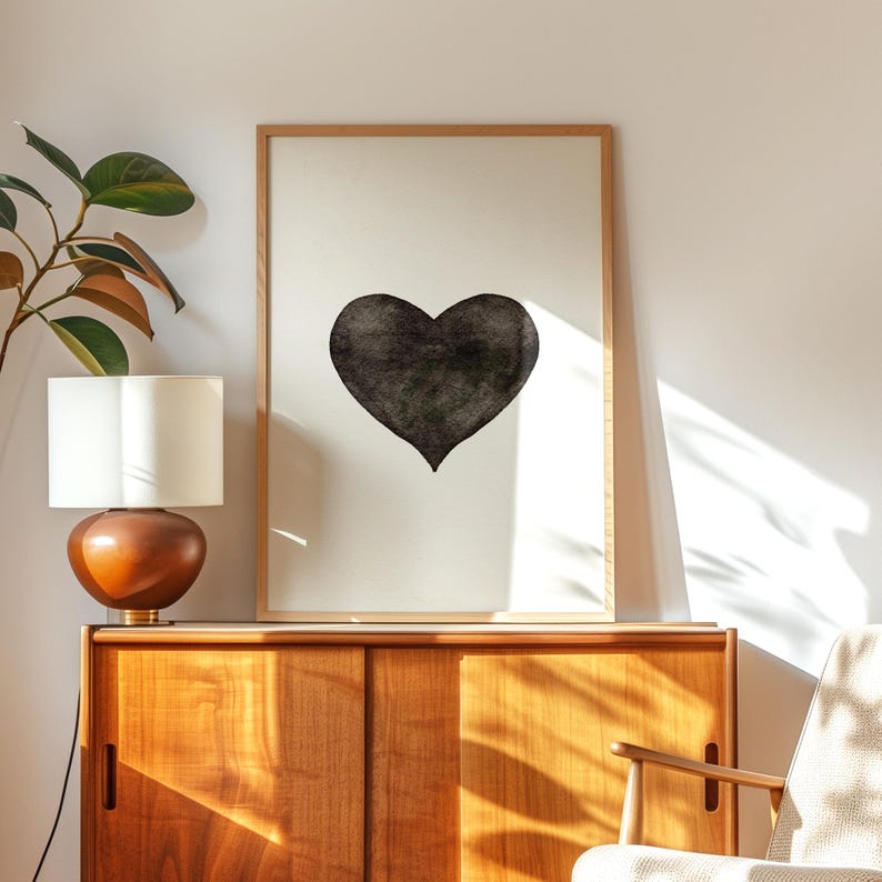 May include: A framed art print featuring a large, textured black heart on a white background. The artwork is displayed on a light wood cabinet, next to a lamp with a white shade and a leafy plant. The room is illuminated by natural light.