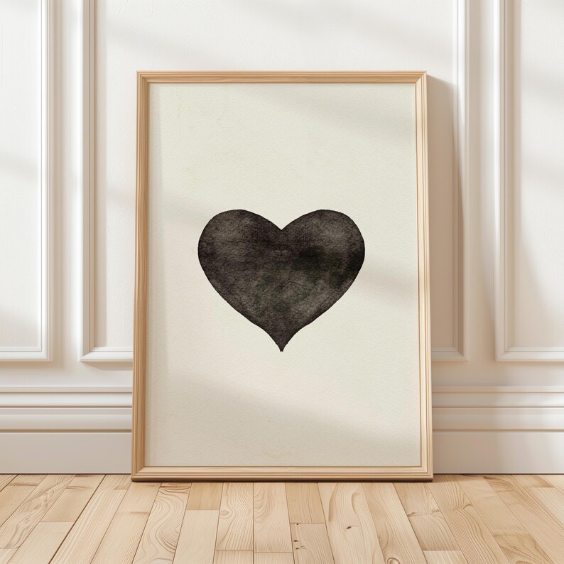 May include: A framed art print featuring a black heart on a cream-colored background. The heart is centrally positioned and has a textured appearance. The frame is made of light-colored wood, and the artwork is displayed against a white wall and wooden floor.