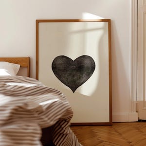 May include: A framed art print featuring a large, textured black heart on a cream-colored background. The artwork is displayed in a room with a bed and striped bedding, creating a cozy, minimalist aesthetic.