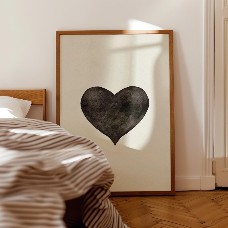 May include: A framed art print featuring a large, textured black heart on a cream-colored background. The artwork is displayed in a room with a bed and striped bedding, creating a cozy, minimalist aesthetic.