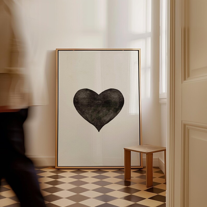 May include: A large framed art print featuring a black heart on a cream background. The artwork is displayed in a room with a small wooden side table and a checkered floor. The frame is gold.