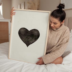 May include: A framed art print featuring a large, textured black heart on a light background. The artwork is encased in a light-colored wooden frame. The print is a minimalist design, suitable for home decor.