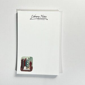 May include: A stack of white notepaper with the words "Library Notes" in black script at the top. A small watercolor illustration of books and foliage is in the lower left corner.