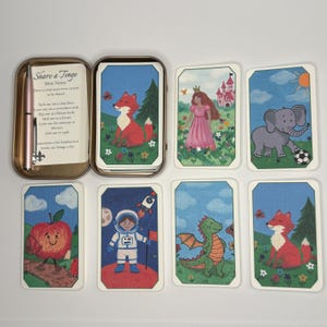 May include: A set of illustrated mini note cards in a metal tin with a pen. The cards feature colorful drawings of a fox, a princess, an elephant, an apple, an astronaut, and a dragon. The tin includes a card with the text Share a Tinge Mini Notes.