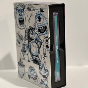 Puede incluir: Una caja negra con una cubierta blanca que presenta bocetos de personajes de la película animada Monsters, Inc. La cubierta tiene el título "Monsters, Inc." impreso en ella. La caja está cerrada y la columna vertebral es visible, mostrando el título "Disney Pixar Monsters, Inc."