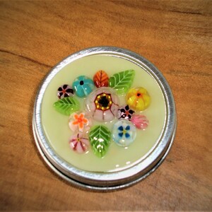 May include: A round, silver-rimmed container with a pale yellow interior. The container is decorated with colorful glass flowers and leaves. The flowers are various colors, including pink, orange, blue, and yellow. The leaves are green.
