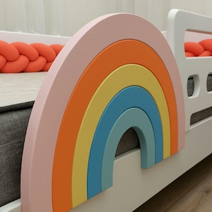 May include: A white wooden bed frame with a rainbow-shaped headboard. The rainbow is painted in shades of pink, orange, yellow, blue, and green.