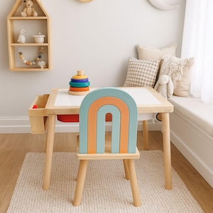 Può includere: Un set di tavolo e sedia per bambini con un design a forma di arcobaleno. Lo schienale della sedia è un arcobaleno arancione e blu. Sono visibili anche una mensola in legno a forma di casa e due decorazioni murali a forma di luna. Un tappeto e dei cuscini completano la scena.