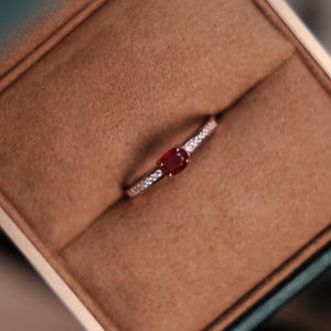 Op de afbeelding: Een delicate ring met een rode ovale edelsteen, geaccentueerd door kleine, heldere stenen langs de band. De ring wordt gepresenteerd in een bruine, met fluweel gevoerde doos met een groene buitenkant. De tekst "KALTE FLAMME" is onderaan zichtbaar.