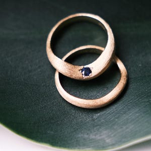 Könnte beinhalten: Zwei goldene Ringe mit gebürsteter Oberfläche, einer davon mit einem kleinen, dunkelblauen Edelstein besetzt. Die Ringe liegen auf einem dunkelgrünen Blatt, was einen Kontrast von Texturen und Farben erzeugt. Verlobungs- oder Eheringe.
