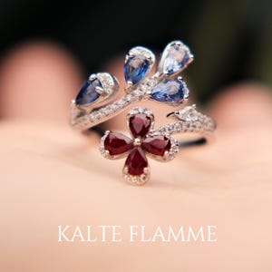 Peut inclure: Bague en argent avec un motif floral orné de pierres précieuses bleues et rouges. La bague présente une fleur rouge à quatre pétales au centre et des pierres bleues en forme de goutte. Le texte "KALTE FLAMME" est en bas.