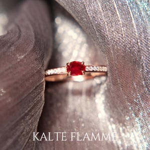 Peut inclure: Une bague en or rose ornée d'une pierre rouge vif au centre, flanquée de petites pierres scintillantes. La bague est mise en valeur sur un tissu argenté chatoyant. Le texte "KALTE FLAMME" est visible.