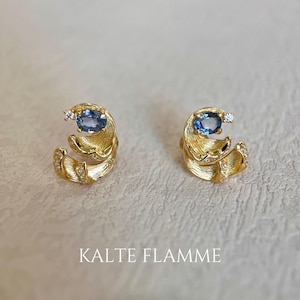 May include: A pair of gold-colored earrings with a unique design. Each earring features a blue oval-shaped gemstone and a small, clear accent stone. The earrings have a textured, swirling pattern and are embellished with small, sparkling details. The text "KALTE FLAMME" is at the bottom.