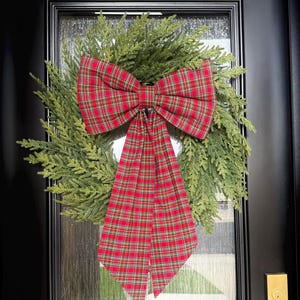May include: A circular wreath made of green cedar-like foliage, featuring a large, prominent bow made of red, green, and yellow plaid fabric. The bow has two long, trailing tails that hang down the center of the wreath, creating a festive appearance.