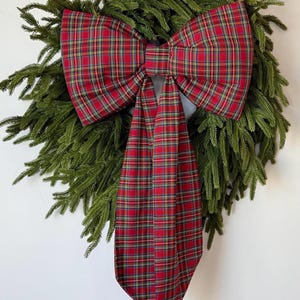 May include: A festive Christmas wreath featuring a lush green base adorned with a large, vibrant red plaid bow. The bow's pattern includes yellow and green accents, adding a touch of holiday cheer to this seasonal decoration.