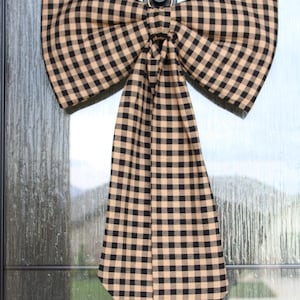 May include: A large, decorative bow made of tan and black checkered fabric, attached to a door. The bow is centered and has a classic shape, with a long, pointed tail hanging down. The fabric has a traditional gingham pattern.