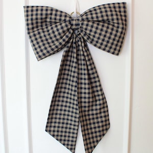 May include: A large decorative bow made of fabric with a navy blue and tan plaid pattern. The bow features two wide loops and two long, hanging tails that taper to points at the bottom. A small gold-toned metal ring is visible at the center knot.
