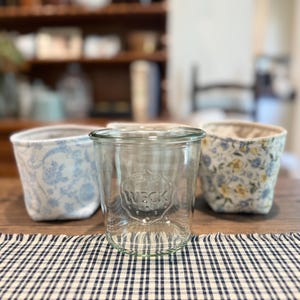 May include: A clear glass jar with the embossed word "WECK" is centered on a wooden table, flanked by two fabric-covered containers. The containers have floral and patterned designs. A checkered tablecloth is in the foreground.