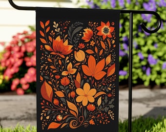 May include: A black garden flag with a colourful floral pattern in orange, rust, and yellow. The flowers and leaves are densely arranged against a black backdrop. The flag is displayed on a black metal garden stake, set in a garden.